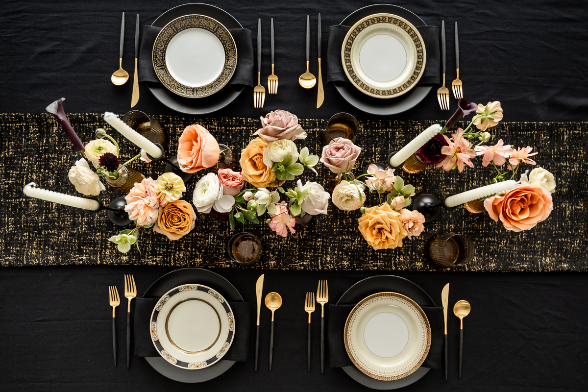 Elegant black and gold table setting with floral arrangements, plates, and black/gold handled cutlery on a black tablecloth.
